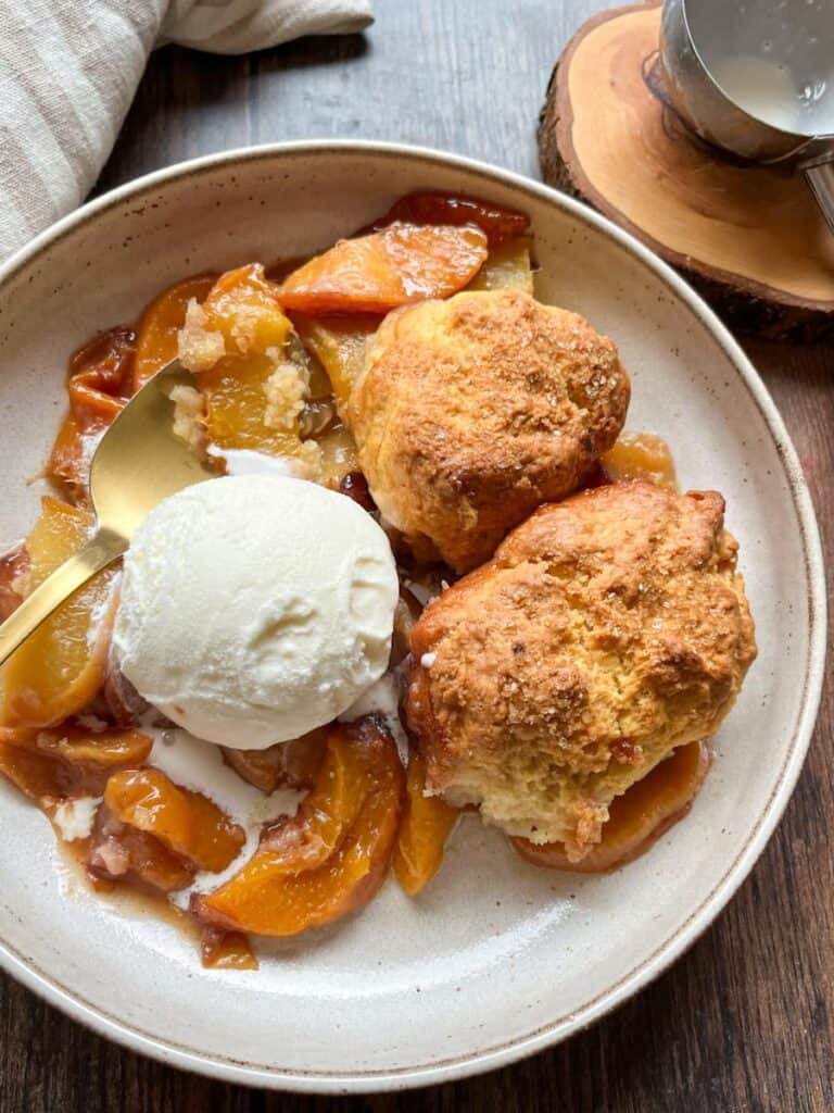 A beige bowl of peach cobbler with a scoop of vanilla ice cream and a gold spoon. A wooden coaster with a silver ice cream scoop resting on it sits in the background.