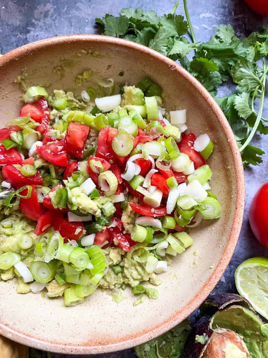 A beige stoneware bowl of mashed avocado, chopped tomatoes and sliced Spring onions.