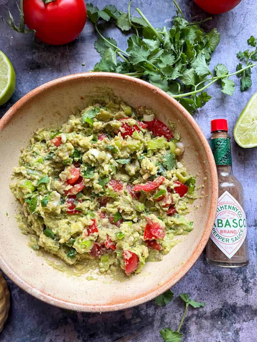 A beige stoneware bowl of mashed ripe avocado, chopped tomatoes and spring onions with a bottle of Tabasco sauce, fresh coriander leaves and lime wedges. 