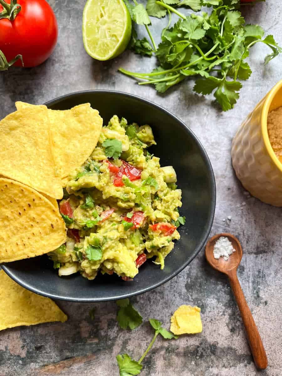 a black bowl of Guacamole with chopped tomatoes or Spring onions with yellow tortilla chips. Fresh coriander and a pot of sea salt with a small wooden spoon sit beside the bowl.