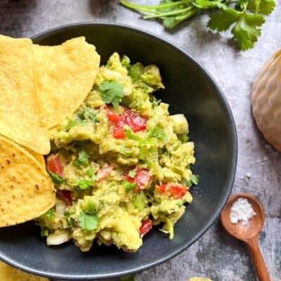 A black bowl of Guacamole with chopped tomatoes and spring onions with yellow tortilla chips resting on the bowl.