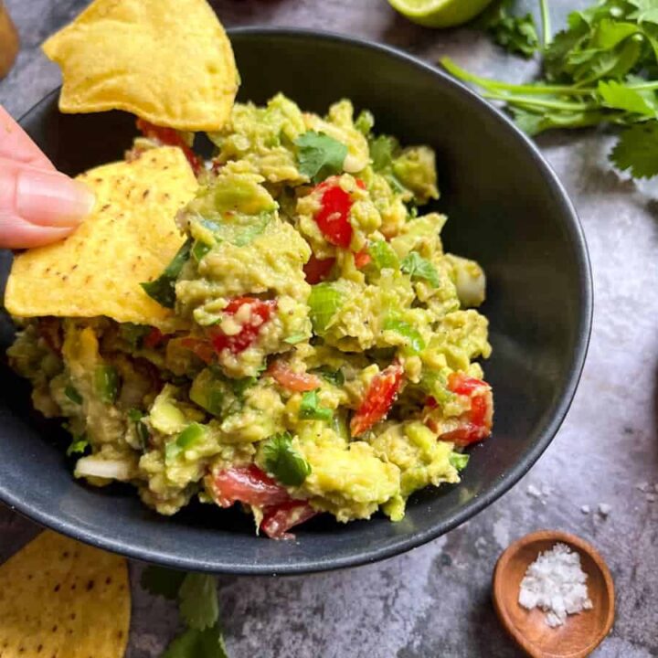 A hand holding a yellow tortilla chip over a black bowl of Guacamole with chopped tomatoes and Spring onions.