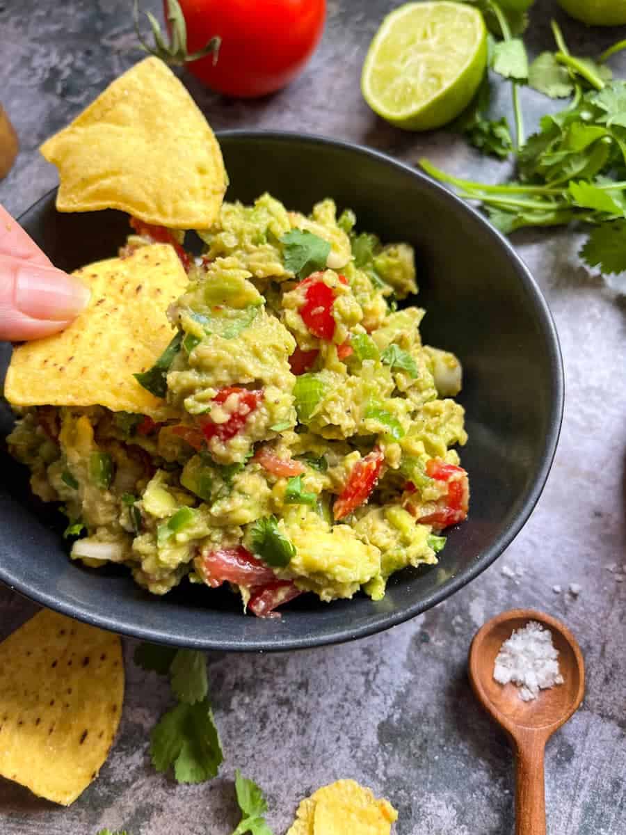 A hand holding a yellow tortilla chip over a black bowl of Guacamole with chopped tomatoes and Spring onions. 