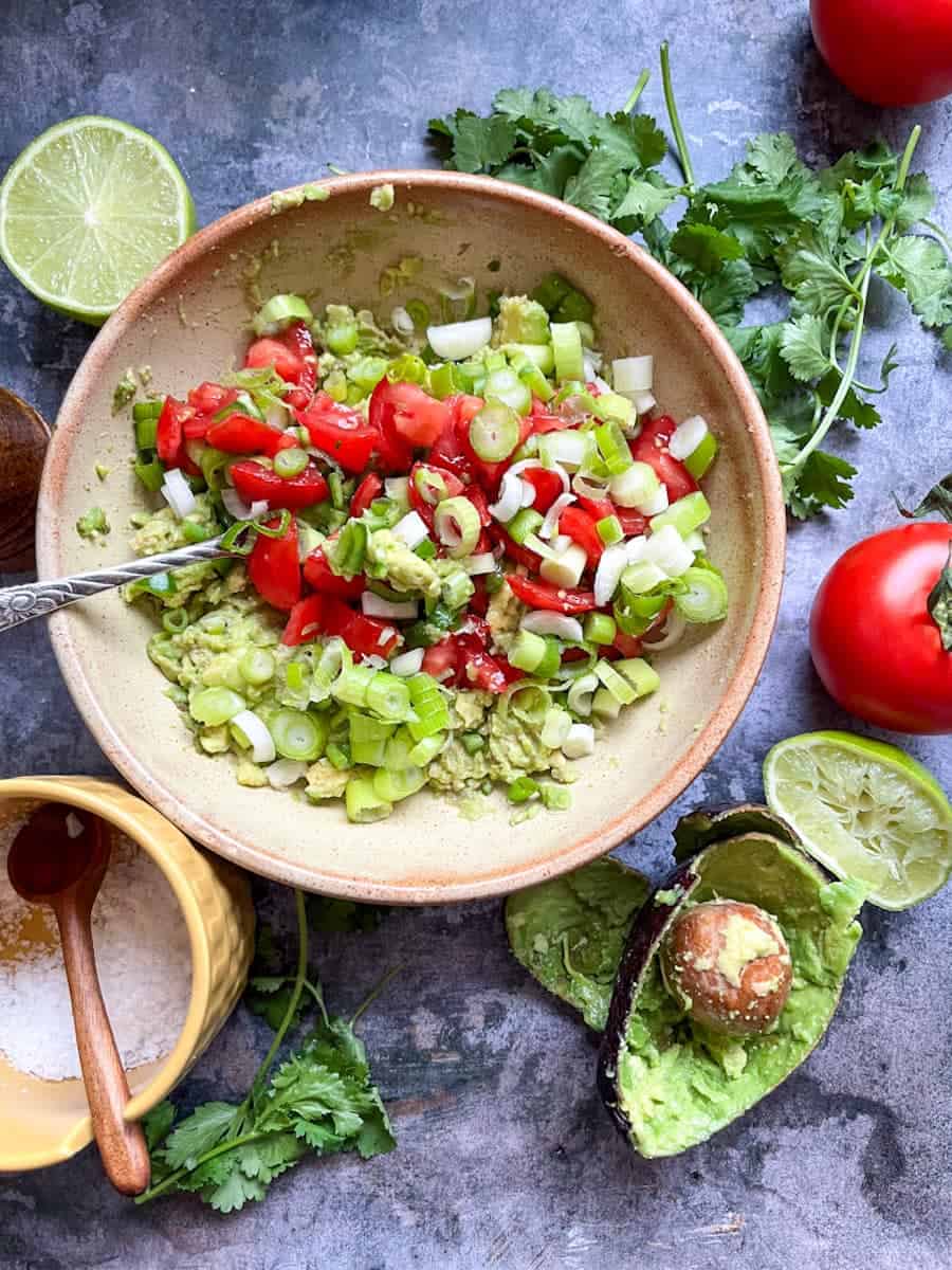 A beige bowl of mashed avocado, chopped tomatoes, chopped spring onions with a fork. A yellow salt pot with a wooden spoon, avocado skins, a large tomato, fresh coriander and a lime wedge surround the bowl.