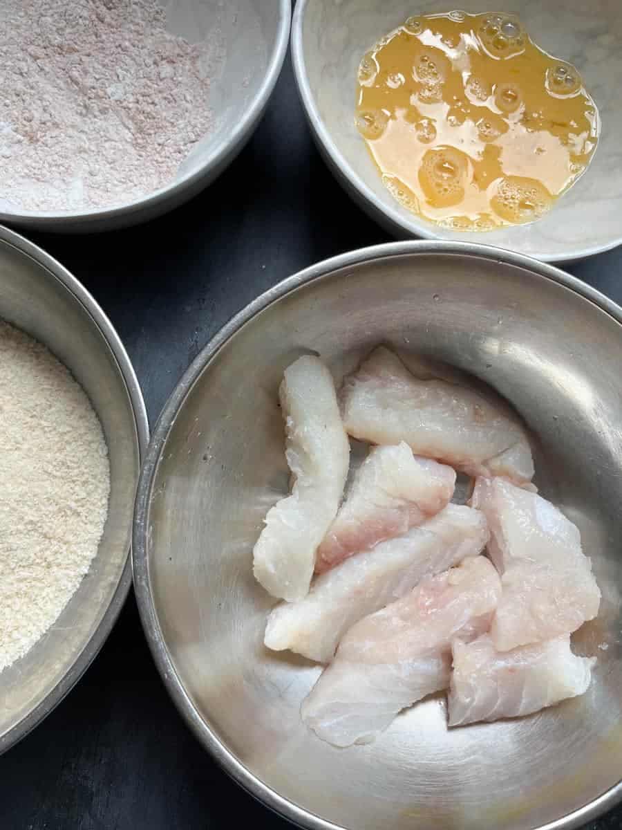 A silver bowl of sliced fish fillets, a white bowl of beaten eggs, a white bowl of flour and a silver bowl of panko breadcrumbs.