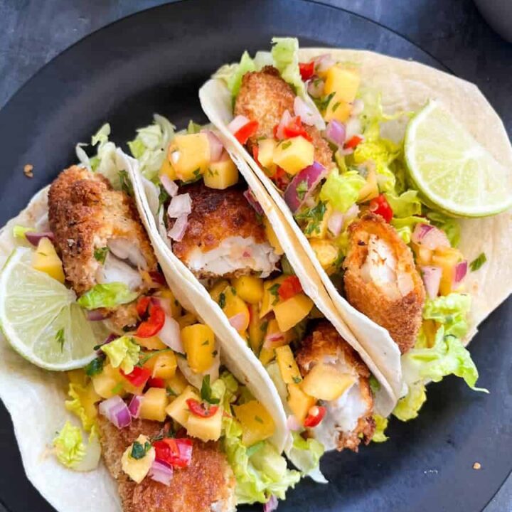 three fish tacos with mango and chilli salad, lettuce and lime wedges on a black plate.