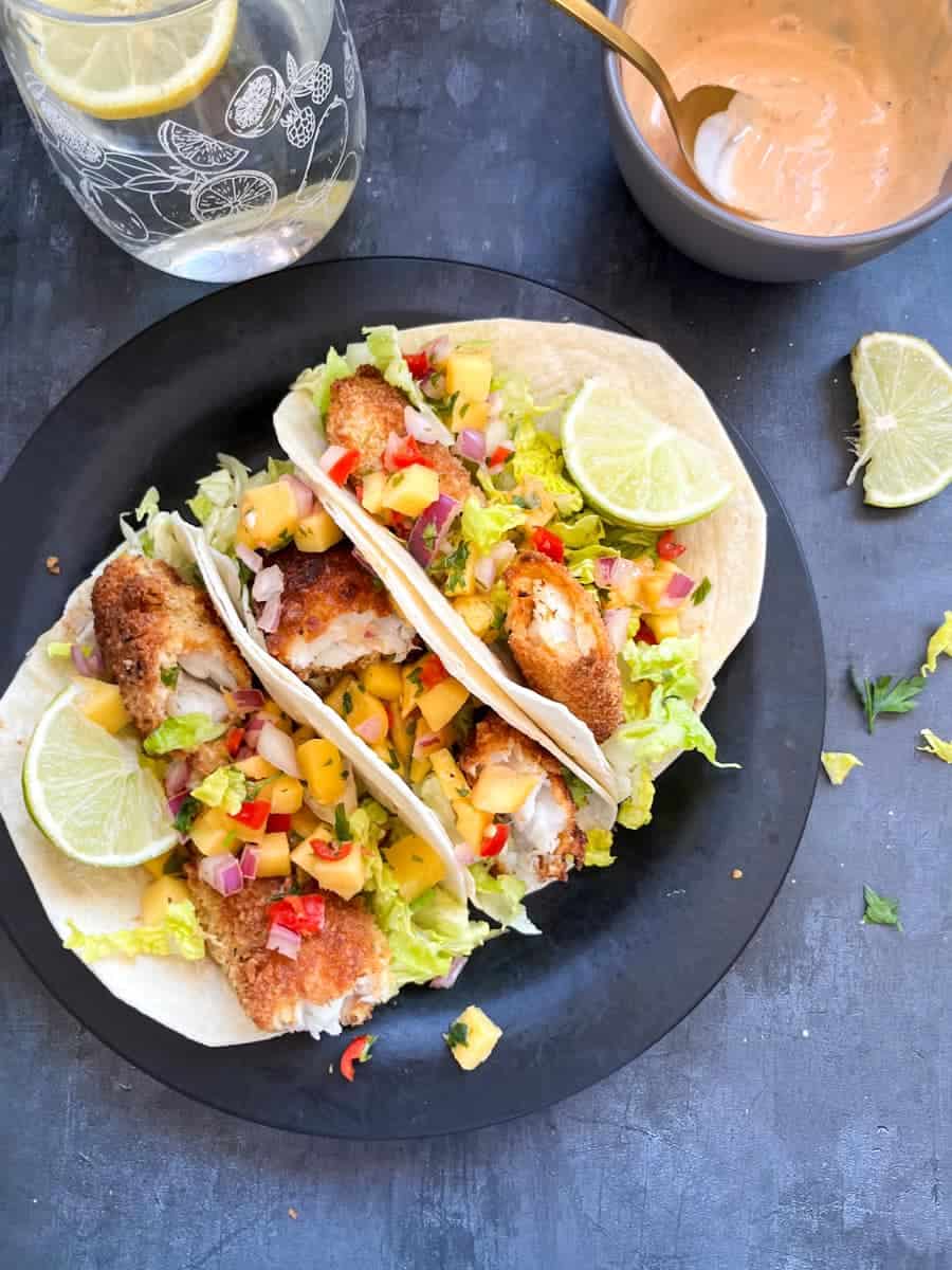 three fish tacos with mango chilli salsa and lime wedges on a black plate with a glass of water with lemon and a grey bowl of chipotle mayonnaise with a spoon.