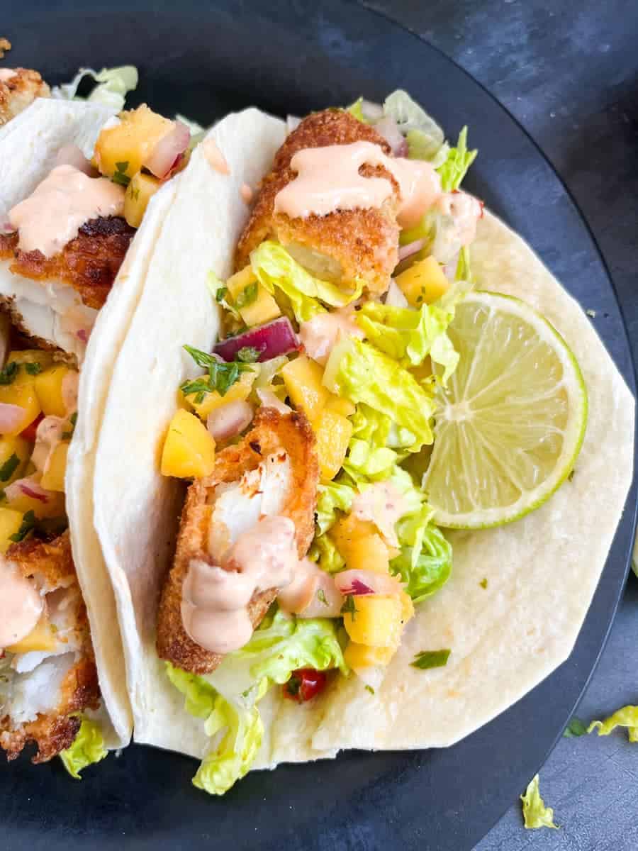 Two fish tacos with breadcrumbed fish fillets with mango, red onion, lettuce and a slice of lime on a black plate.