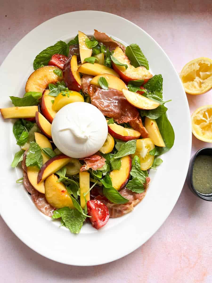 A peach salad with green salad leaves, fresh herbs, grilled prosciutto, tomatoes and a ball of Burrata cheese on a white plate.