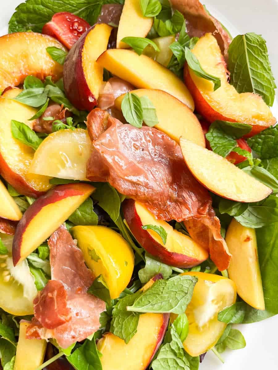 A close up photo of a green salad with sliced peaches, grilled prosciutto and sliced yellow and red tomatoes.