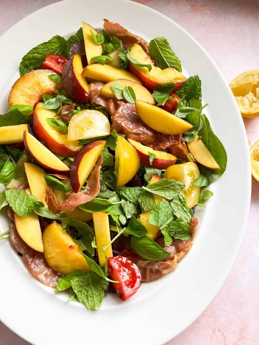 A salad with green leaves, fresh mint and basil, sliced peaches and sliced red and yellow tomatoes on a white plate.