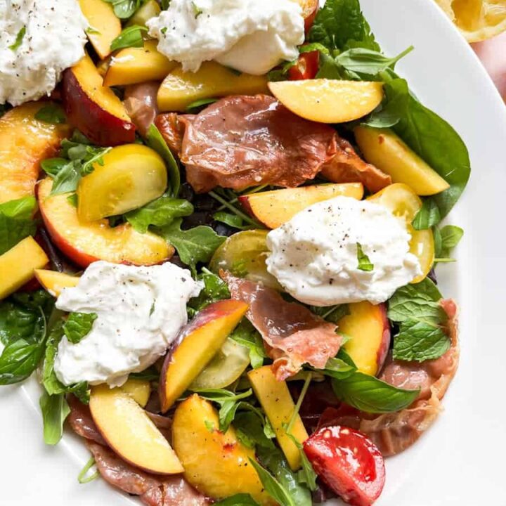A green salad with sliced peaches, grilled proscuitto, red and yellow tomatoes and Burrata cheese on a white plate.