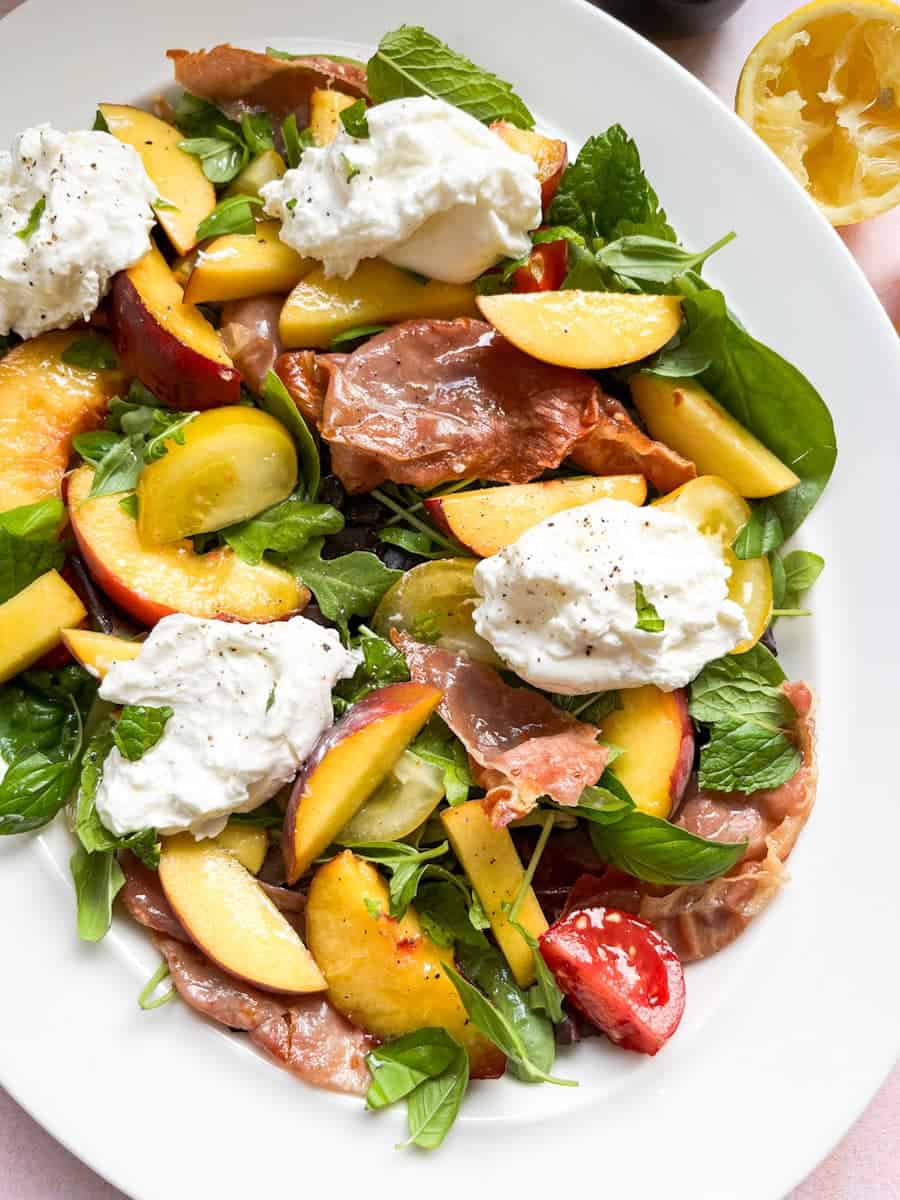 A green salad with sliced peaches, grilled proscuitto, red and yellow tomatoes and Burrata cheese on a white plate.