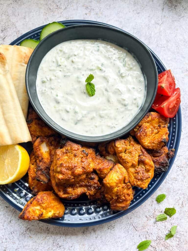 A blue plate of cooked spiced chicken pieces, sliced tomatoes, sliced cucumber, pitta bread, a halved lemon and a black bowl of Tzatziki dip.