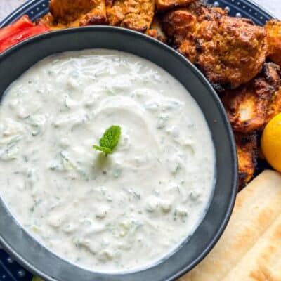 A black bowl of Tzatziki dip with cooked spiced chicken pieces, sliced cucumber and pitta bread.