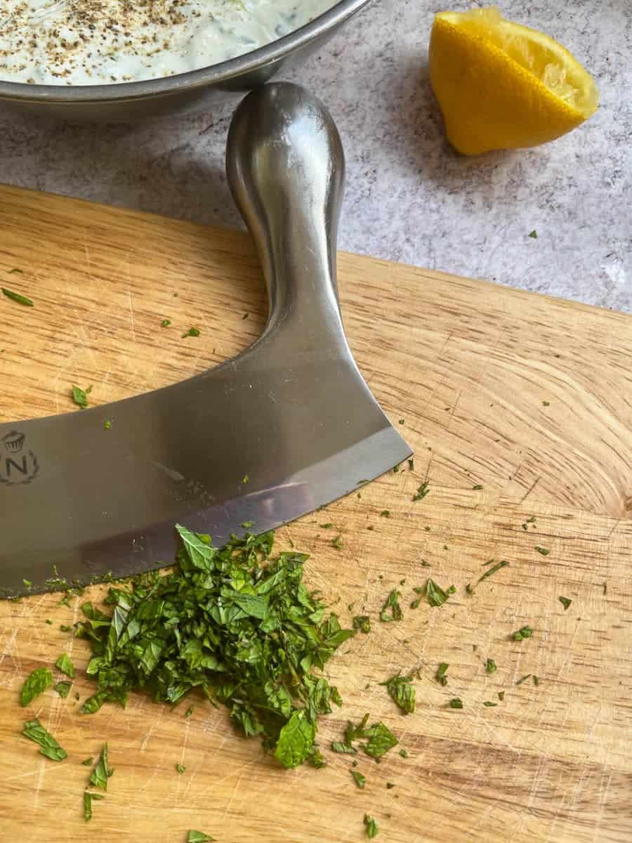 A wooden chopping board with a silver mezzaluna and freshly chopped mint, a squeezed lemon half and a silver bowl of Greek yoghurt seasoned with black pepper.