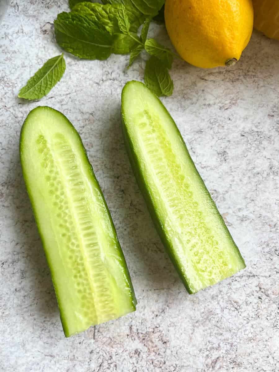 A cucumber sliced in half, a lemon and a small bunch of fresh mint on a grey and white backdrop.