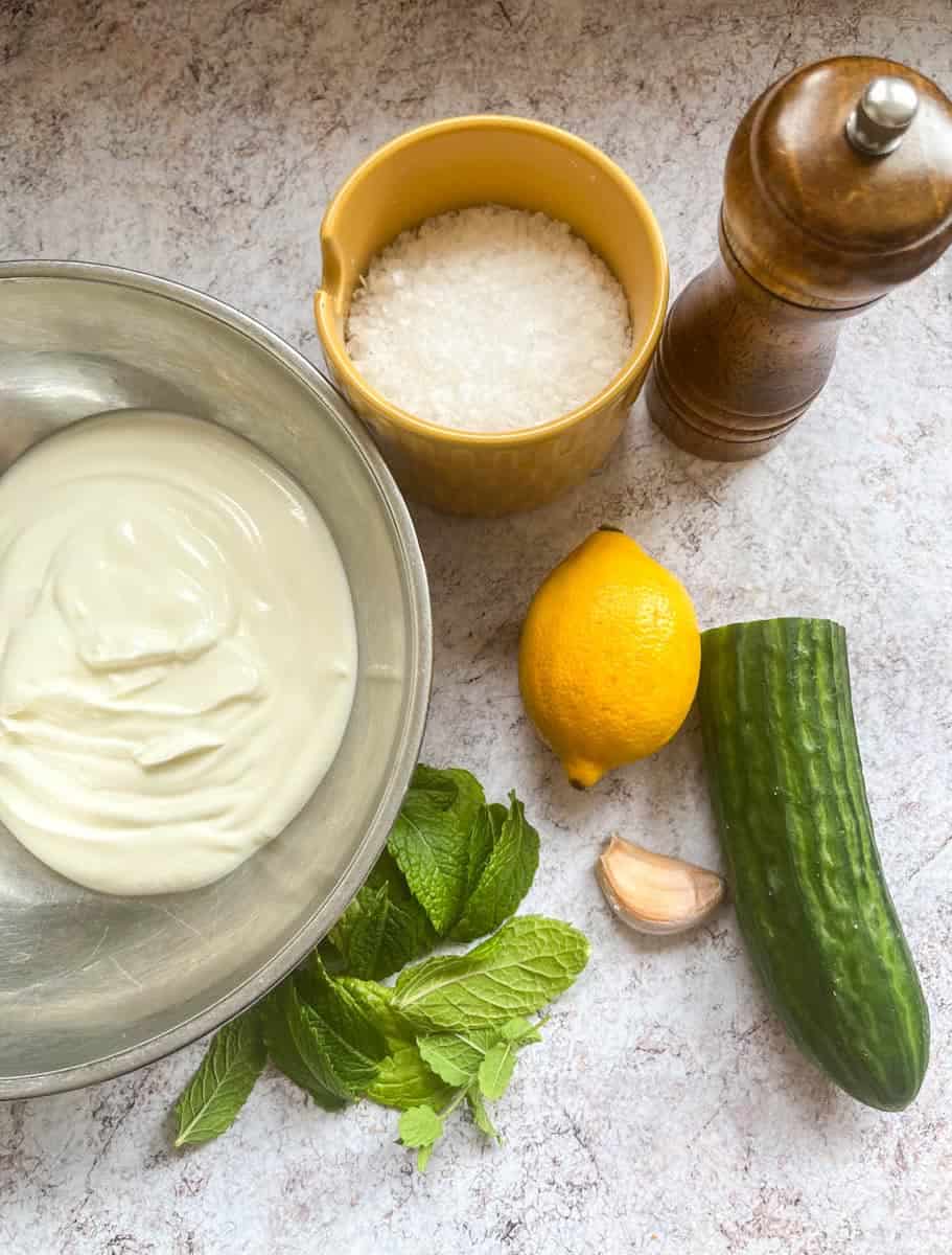 A silver bowl of Greek yoghurt, a yellow pot of flaky sea salt, a wooden pepper mill, half a cucumber, a lemon, a garlic clove and a bunch of fresh mint on a grey and white backdrop.