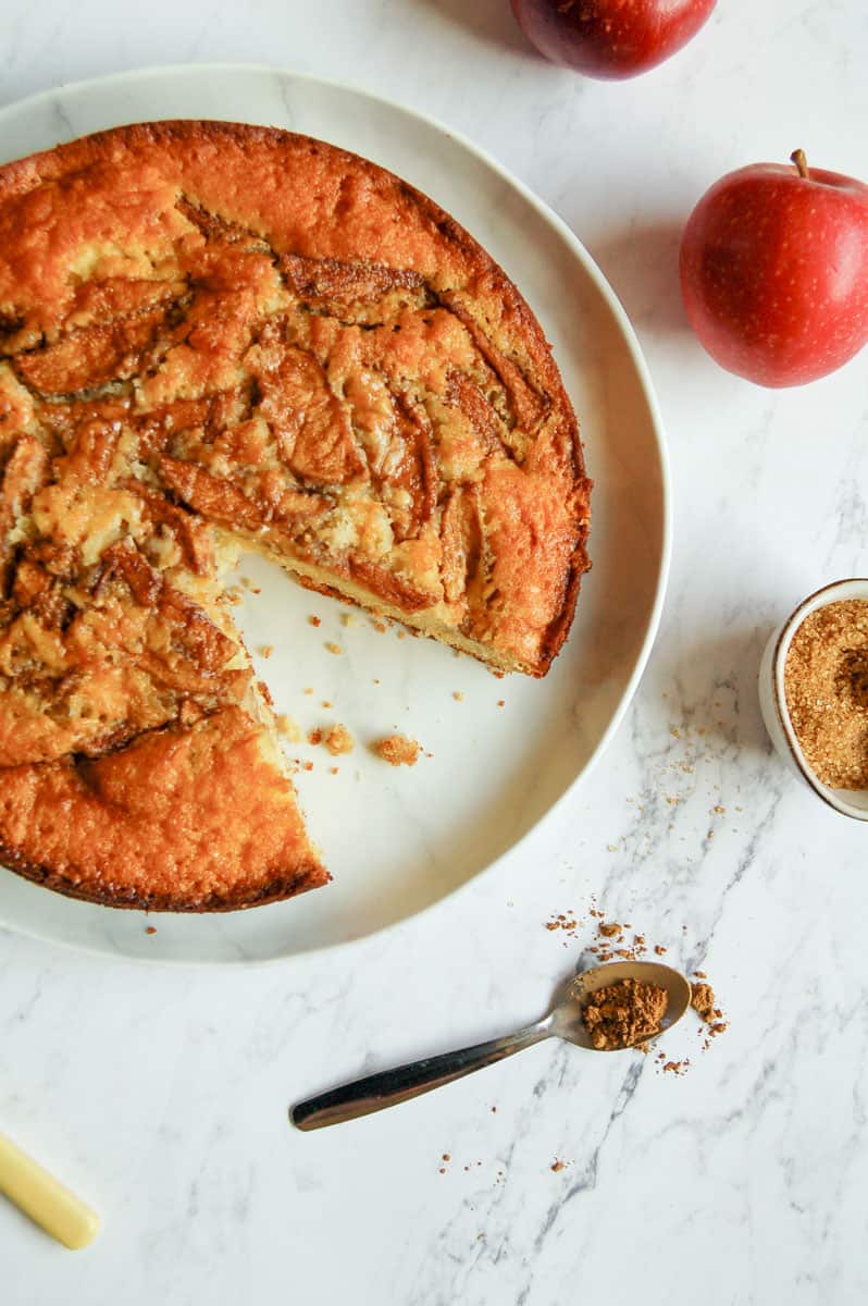 An apple cake with a slice remove on a white plate, a teaspoon of ground cinnamon, a small pot of brown sugar and two red apples.
