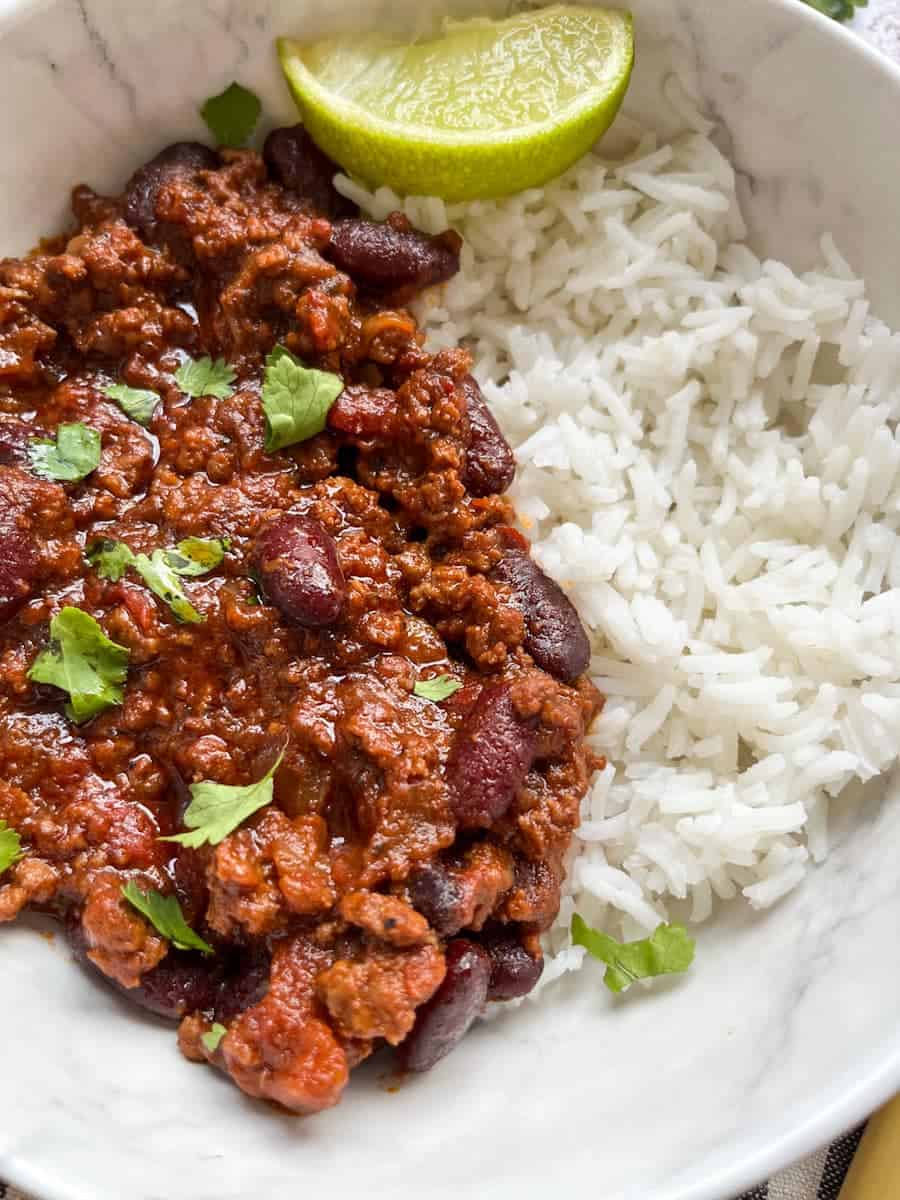 a white bowl of minced beef chilli con carne with rice and a lime wedge.