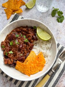 A white bowl of minced beef chilli con carne with rice, tortilla chips and a lime wedge.
