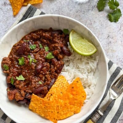 A white bowl of minced beef chilli con carne with rice, tortilla chips and a lime wedge.