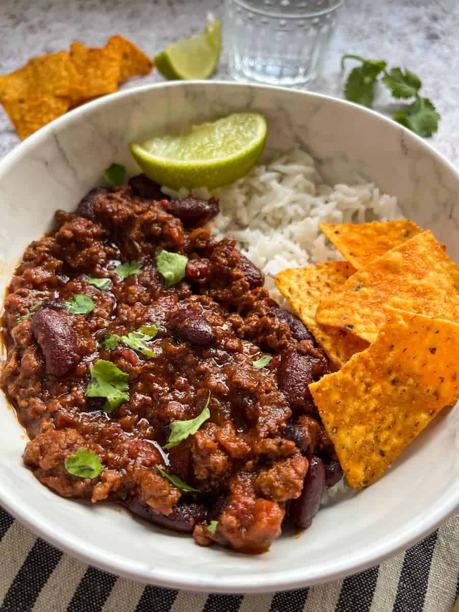 A white bowl of minced beef chilli sprinkled with fresh coriander with rice, tortilla chips and a lime wedge.