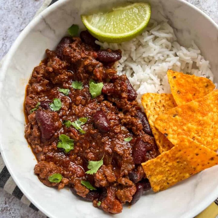 a white bowl of minced beef chilli with rice, tortilla chips and a lime wedge sitting on a striped tea towel.