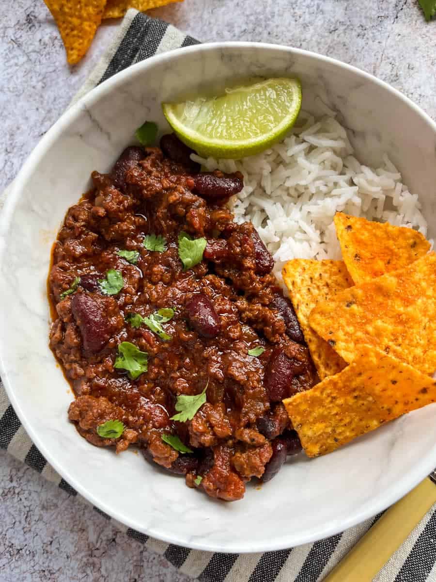 a white bowl of minced beef chilli with rice, tortilla chips and a lime wedge sitting on a striped tea towel.