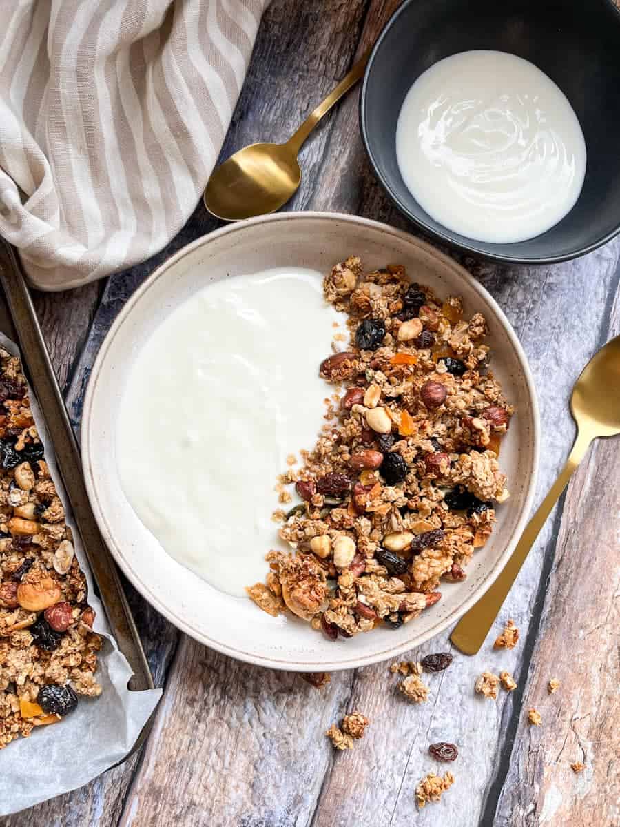 A beige bowl of granola with plain yoghurt, a black bowl of yoghurt, two gold spoons a a beige and white striped tea towel.