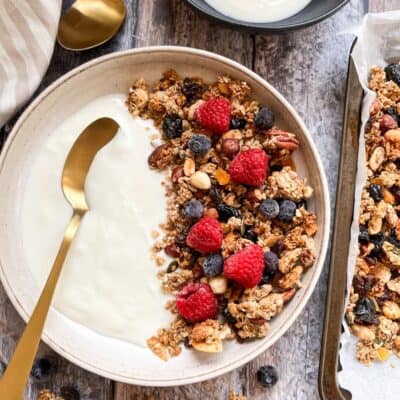 A beige bowl of granola with plain yoghurt, raspberries and blueberries and a gold spoon, a black bowl of yoghurt and a tray of granola.
