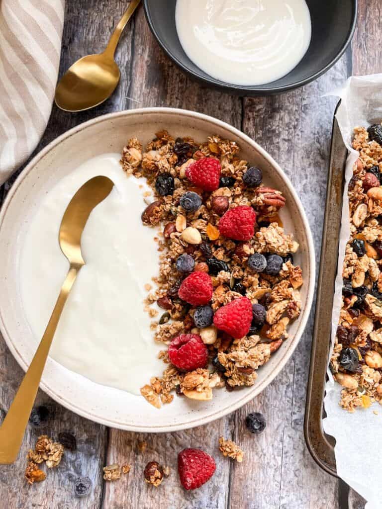A beige bowl of granola with plain yoghurt, raspberries and blueberries and a gold spoon, a black bowl of yoghurt and a tray of granola.