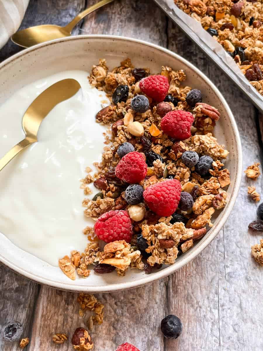 A beige bowl of granola with nuts, seeds and dried apricots and cherries with plain yoghurt, raspberries, blueberries and a gold spoon.