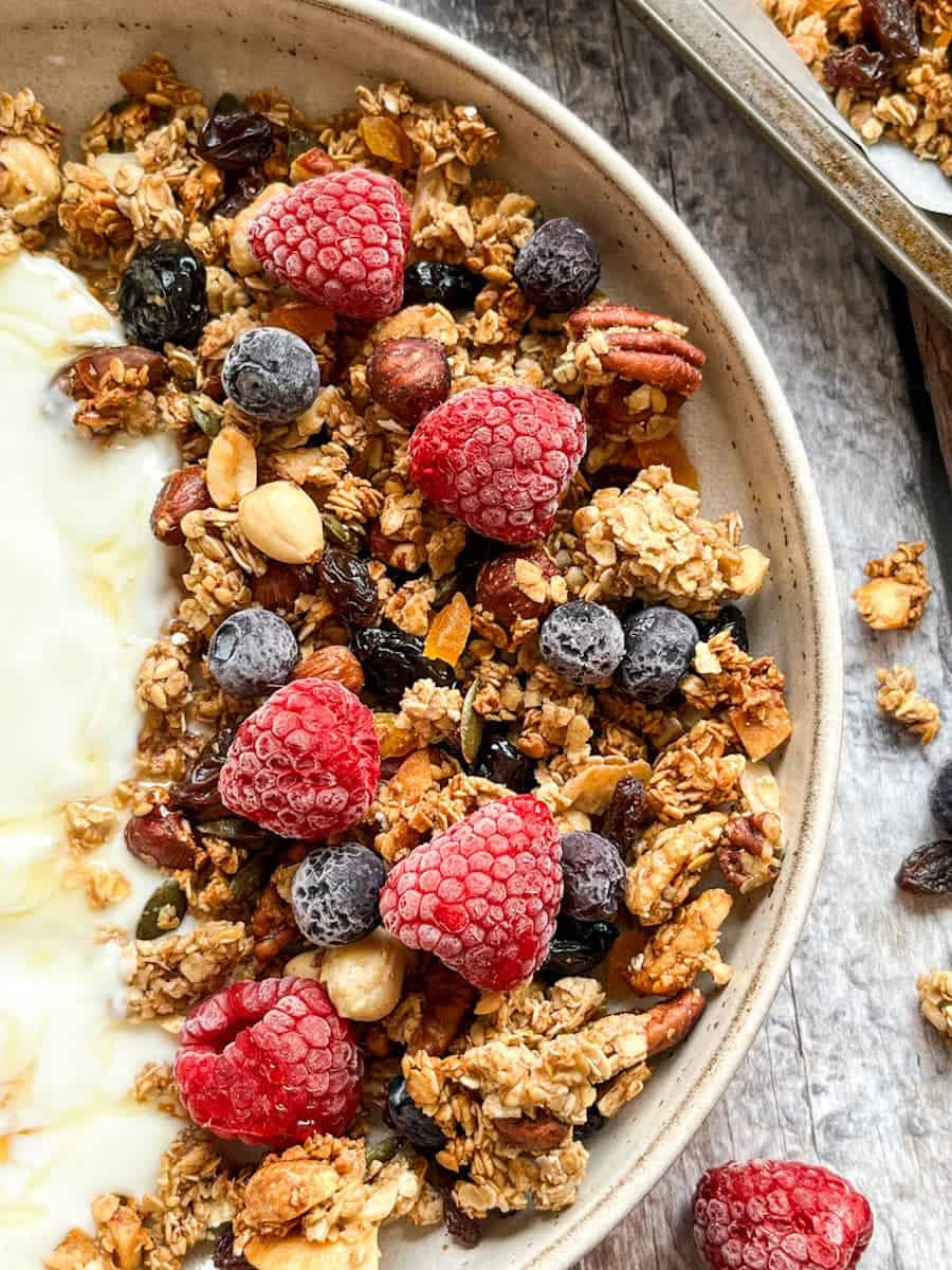 A beige stoneware bowl of air fryer granola with dried apricots, cherries, raspberries and blueberries and plain yoghurt.