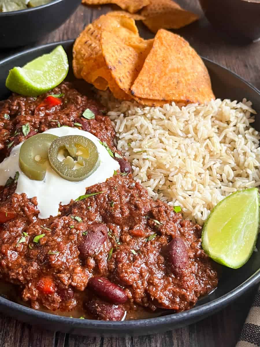 A black stoneware bowl of beef chilli with kidney beans and rice topped with sour cream, green jalepenos, tortilla chips and lime wedges.