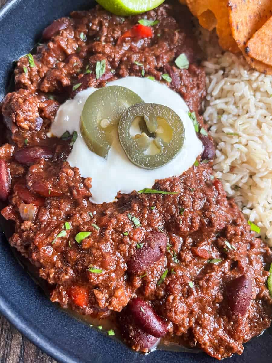 A bowl of chilli con carne and rice topped with sour cream and green Jalepenos.