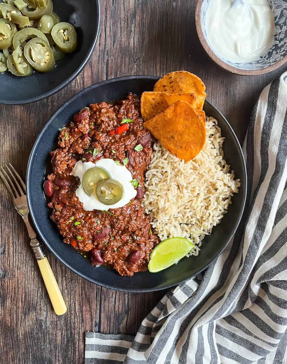 A black stoneware bowl of chilli con carne topped with sour cream, green jalepenos, rice, tortilla chips and a lime wedge. Bowls of sour cream, jalepenos, a striped tea towel and a yellow handled fork are beside the bowl.