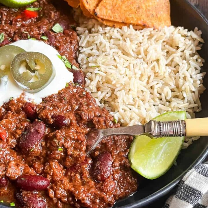 A black stoneware dish of Chilli Con Carne with sour cream, green jalepenos, rice, lime wedges and tortilla chips.