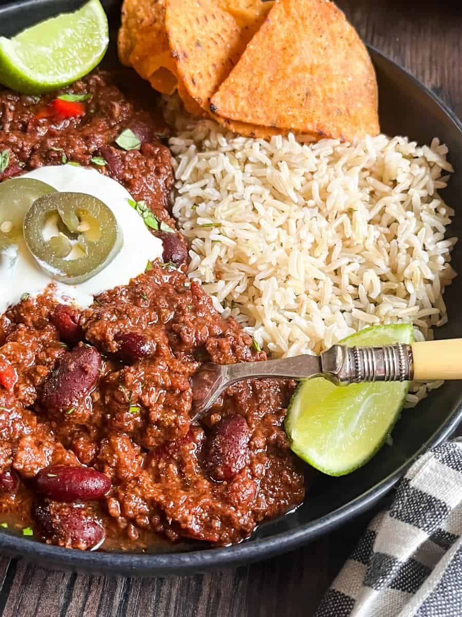 A black stoneware dish of Chilli Con Carne with sour cream, green jalepenos, rice, lime wedges and tortilla chips.