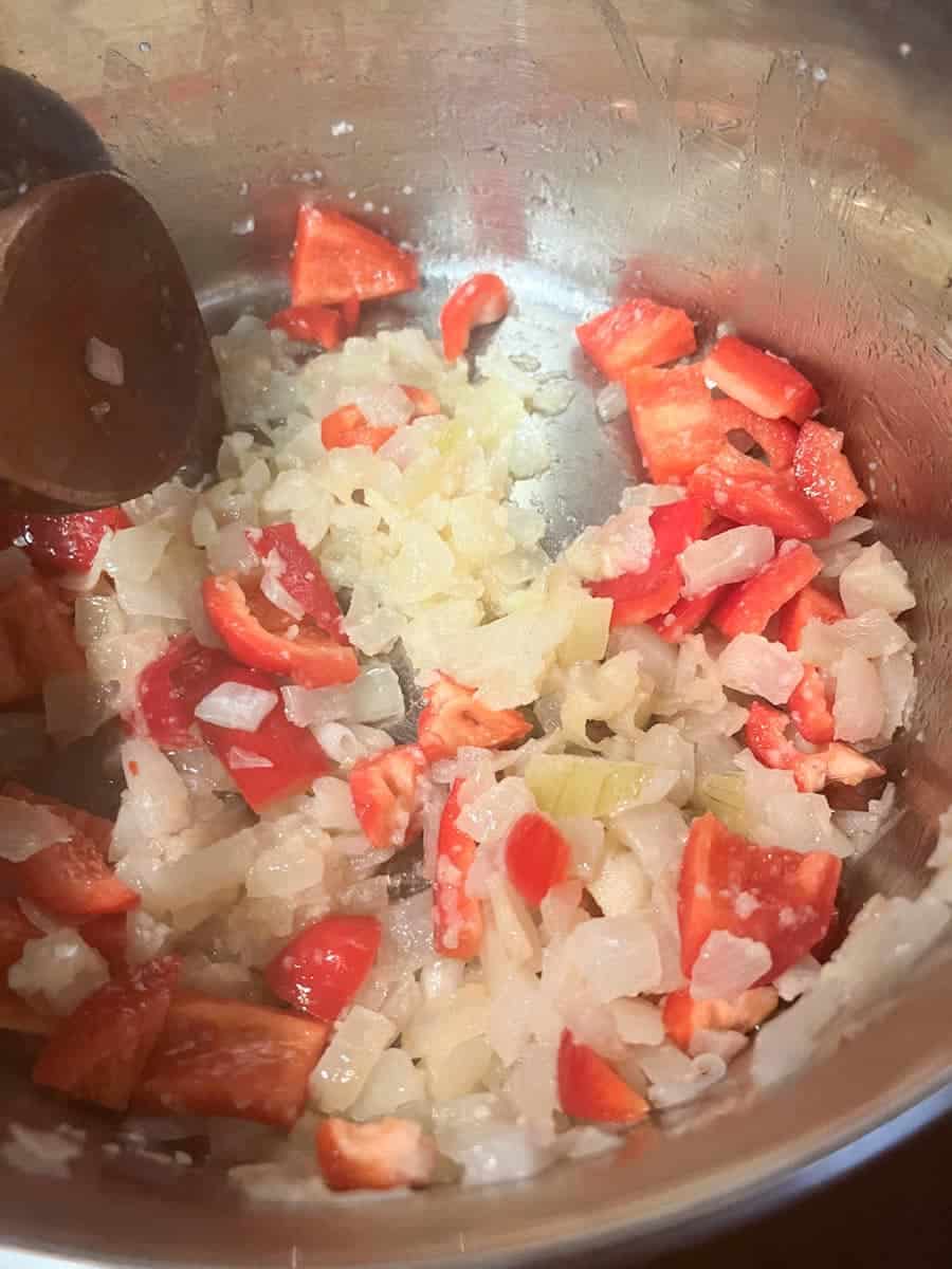 Chopped onion, garlic and diced red peppers cooking in a silver saucepan.