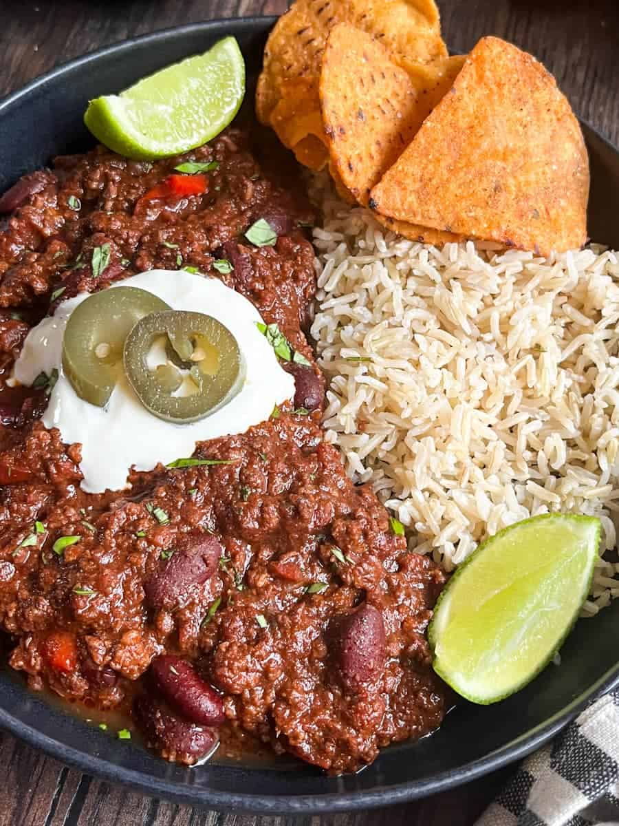 A black bowl of beef chilli with rice, sour cream, jalepenos, tortilla chips and lime wedges.