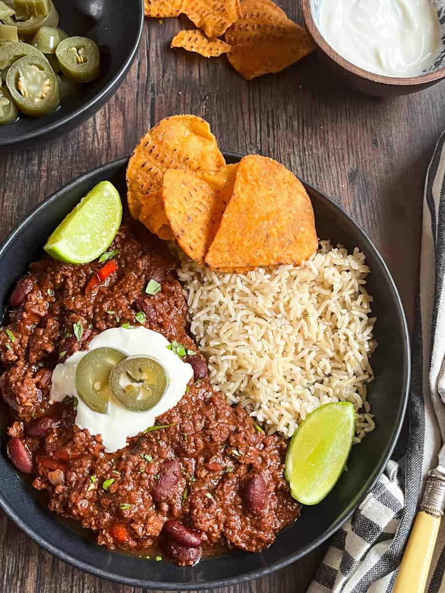 A black bowl of chilli con carne with rice, sour cream, green jalepenos, tortilla chips and lime wedges. Bowls of sour cream, jalepenos and tortilla chips can be partially seen.