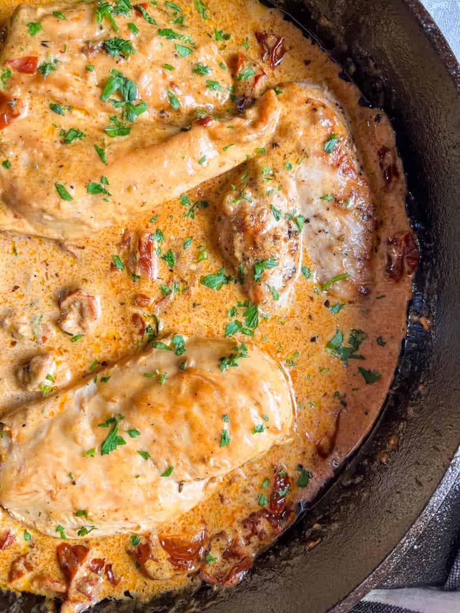 chicken breasts in a creamy sun dried tomato sauce scattered with chopped parsley in a black skillet.