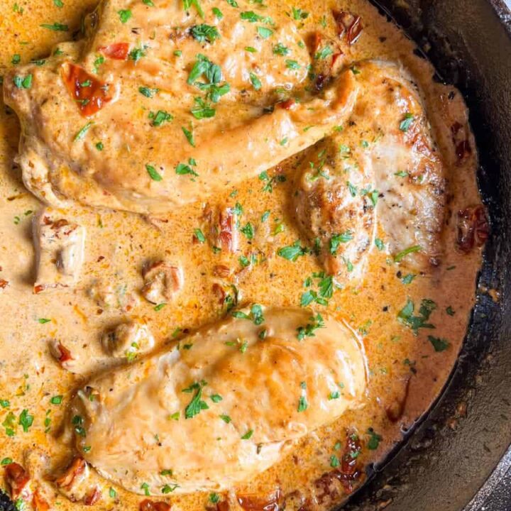 three chicken breasts in a sun dried tomato cream sauce with chopped parsley in a black skillet.