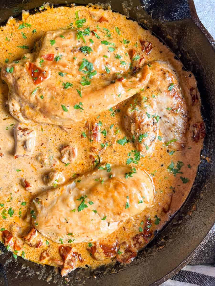 three chicken breasts in a sun dried tomato cream sauce with chopped parsley in a black skillet.