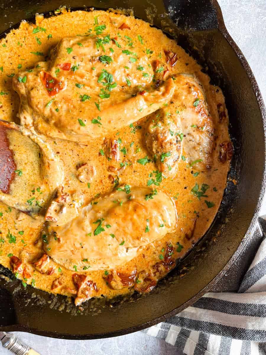 Chicken in a creamy sun dried tomato sauce with chopped parsley in a black skillet.