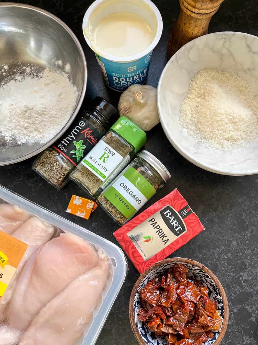 A silver bowl of flour, a white bowl of grated parmesan cheese, a pot of cream, a packet of chicken breasts, a bowl of sun dried tomatoes, a bulb of garlic and pots of dried herbs.