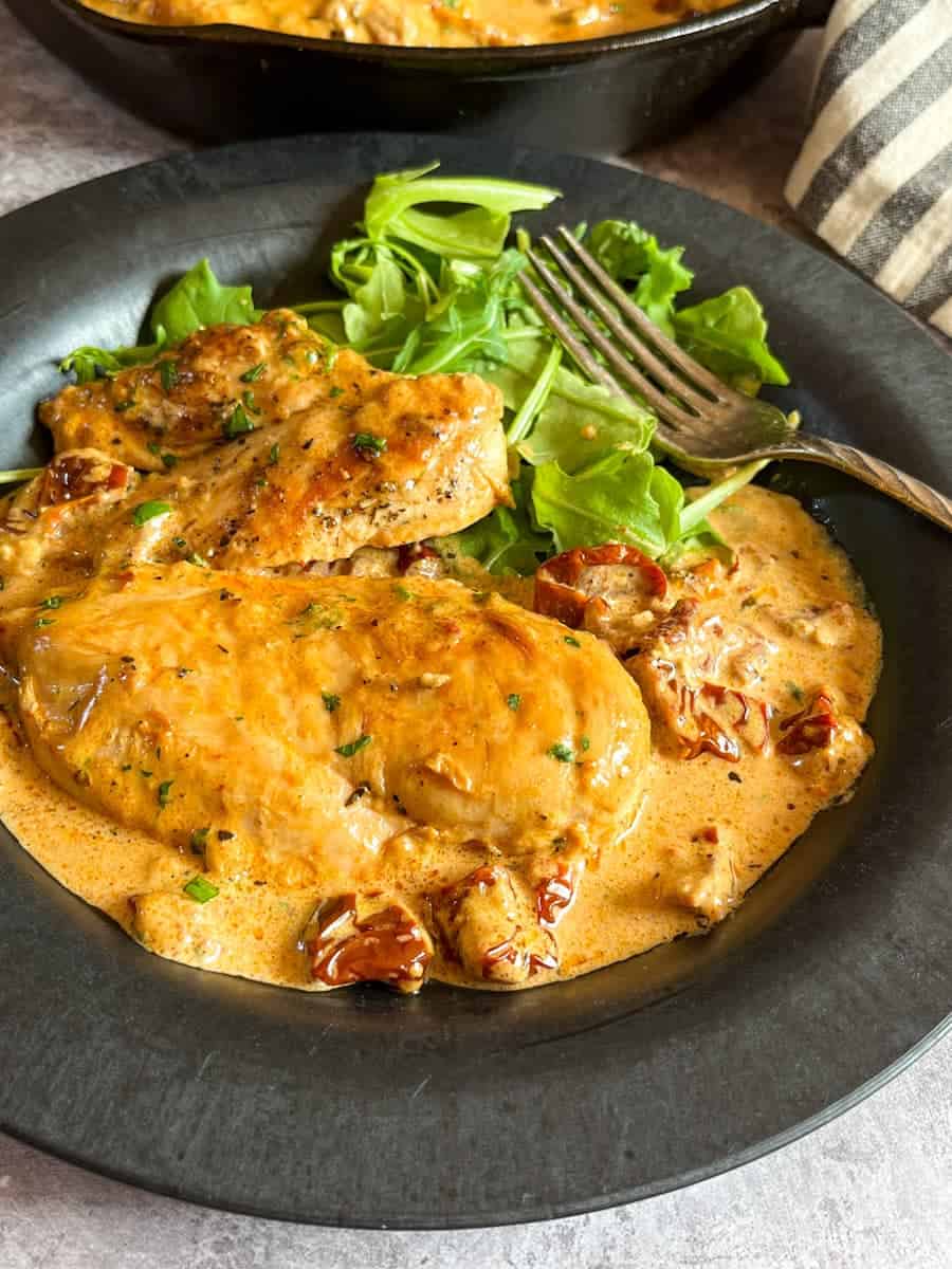 chicken breasts in a cream sun dried tomato sauce with a rocket salad on a black plate with a silver fork.