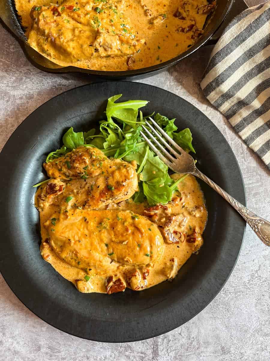 two chicken breasts in a creamy sun dried tomato sauce and a rocket salad on a black plate with a silver fork.