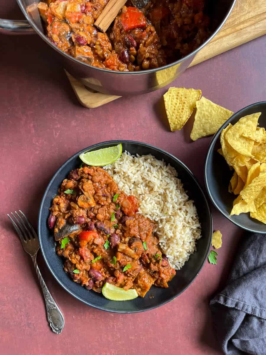 A black bowl of aubergine and lentil chilli with rice, lime wedges, a large silver pot of chilli and a bowl of tortilla chips.